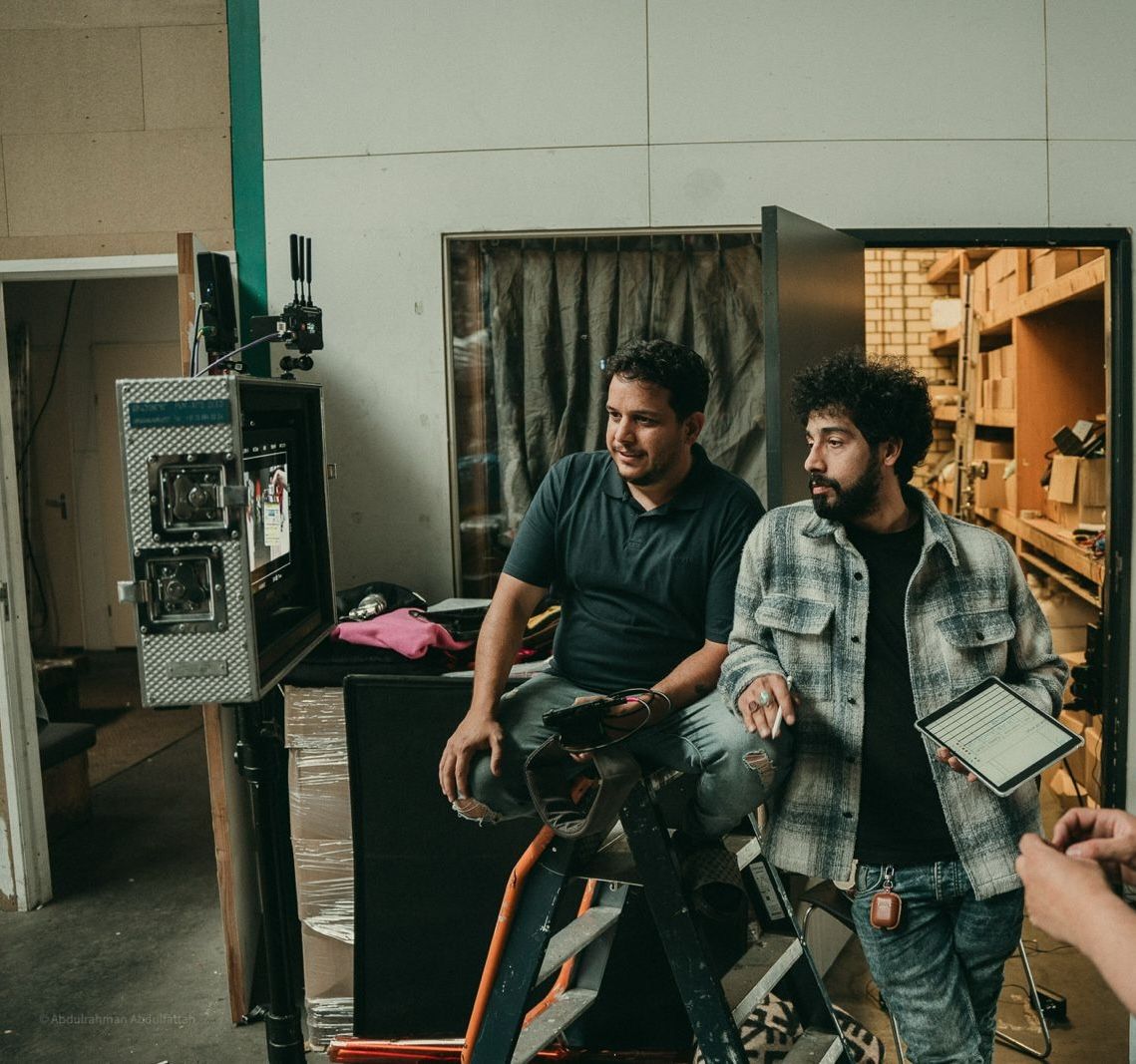 Mohammad Qahees sits with a crew member beside a production monitor on a film set.
